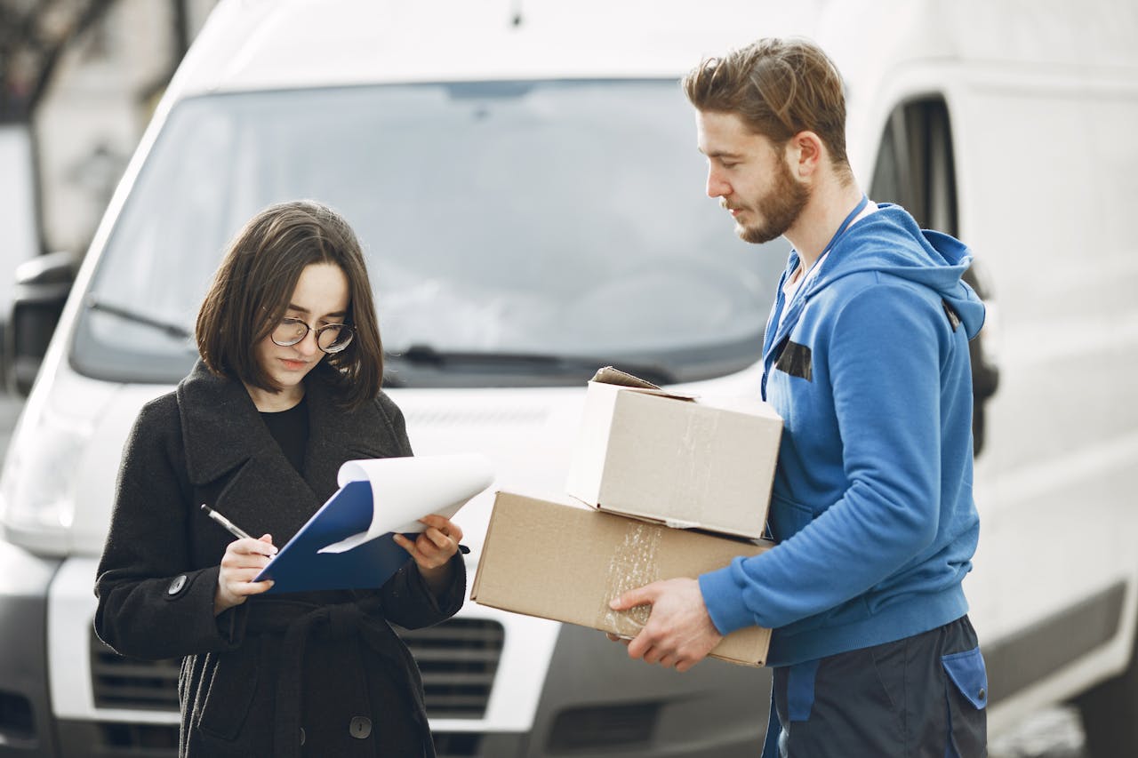 services-01 A courier in blue hands packages to a woman signing documents in front of a delivery van.